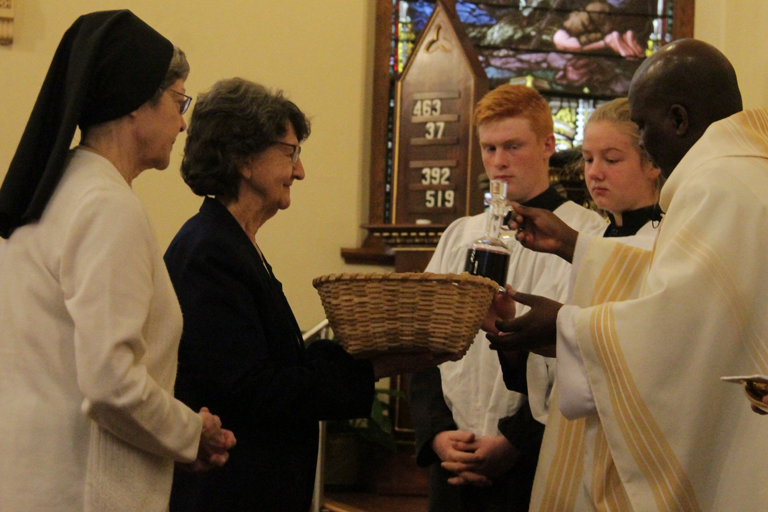 Sister Kathleen Donovan of the School Sisters of Notre Dame, a former principal of St. Thomas the Apostle School, and Sister Rosemary Boessen of the Sisters of Mercy, a St. Thomas native, present the gifts of bread and wine to Father Leonard Mukiibi, administrator of St. Thomas the Apostle parish, during a Mass for the parish’s 150th anniversary.